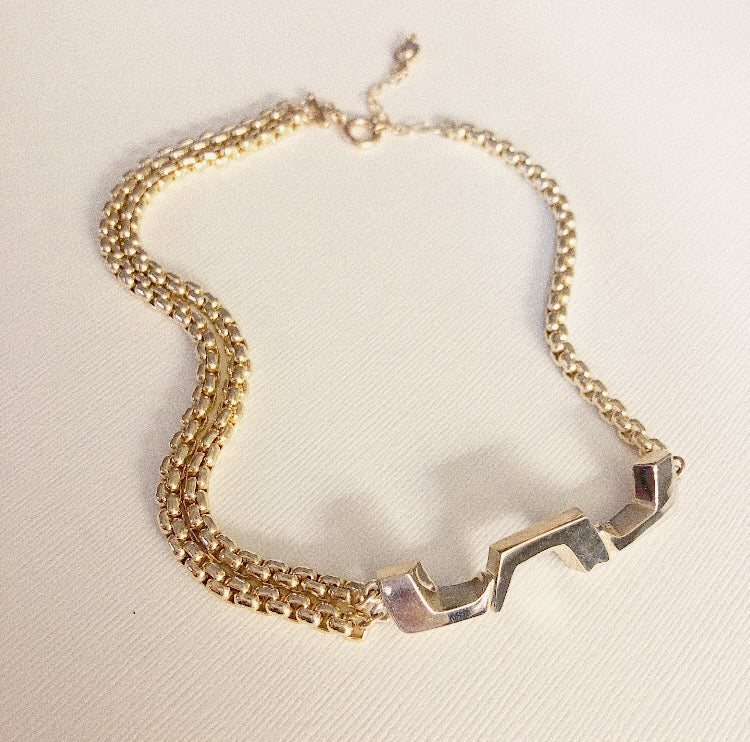 A gold-colored choker necklace with a sculptural pendant and two Venetian chains on the right side, displayed on a white background.